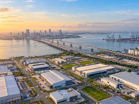 Aerial view of an industrial park in Johor beside the strait, with a bridge and rail line crossing to Singapore and a container port in the distance